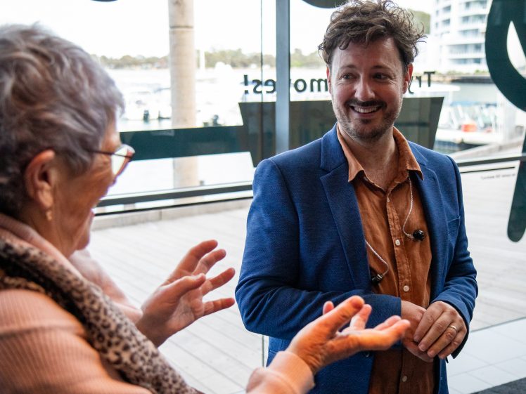 Image of A man in a blue blazer and tan shirt smiles whilst talking with an older woman inside a modern building with large windows overlooking a waterfront. The woman gestures animatedly with her hands.