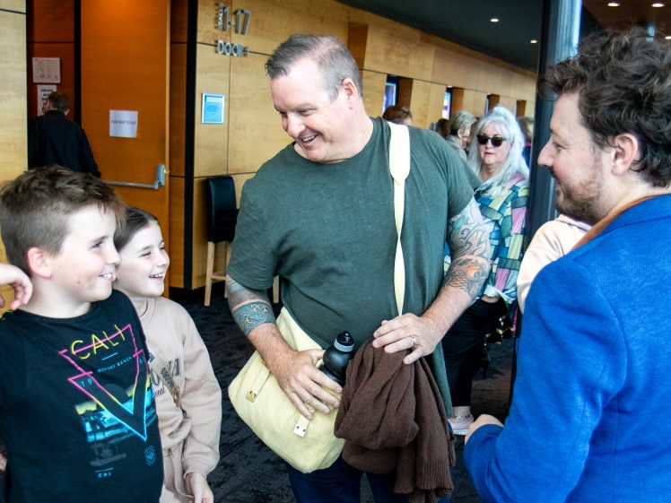 Image of A man with a satchel smiles at two children, while another man in a blue jacket chats with them. They are indoors, with several people in the background near wooden walls and doors.