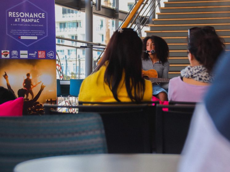 Image of A woman with curly hair plays guitar and sings into a microphone in front of an audience at an indoor event, with a Resonance at MANPAC event banner beside her and a staircase in the background.