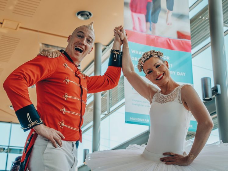Image of A smiling man in a red circus uniform and a woman in a white ballet tutu hold hands and pose joyfully indoors, with large windows and a colourful poster in the background.