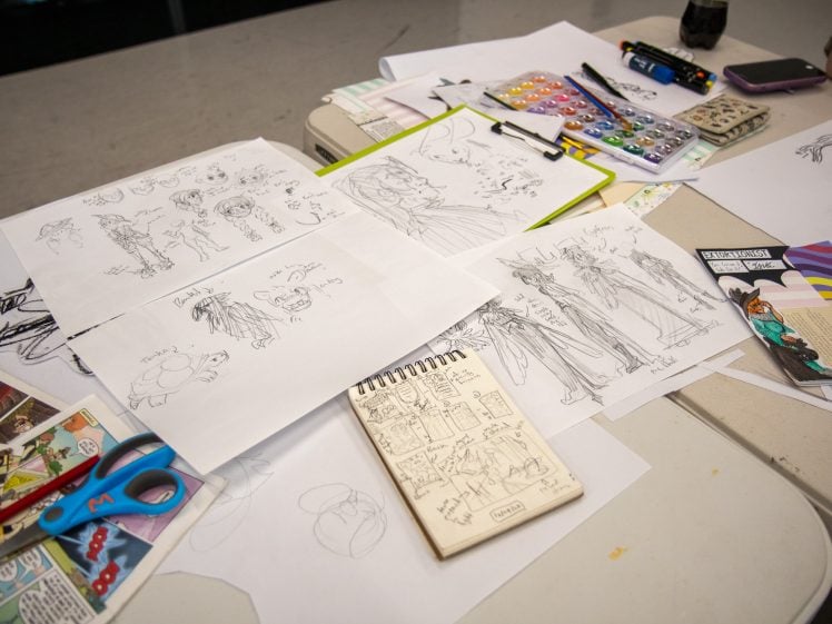 Image of A tabletop covered with character sketches, art supplies, a comic strip, coloured markers, scissors, and watercolour paints. Sheets of paper display various hand-drawn fantasy and cartoon figures.
