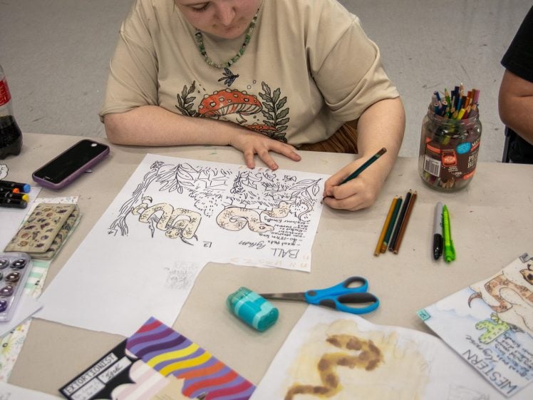 Image of A person with fair hair sits at a table drawing and colouring on paper. Art supplies, including coloured pencils, markers, scissors, and sketchbooks, are scattered on the table.