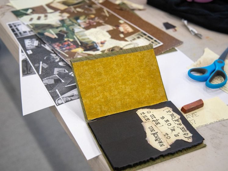 Image of An open handmade journal with yellow and black pages sits on a table surrounded by craft supplies, including scissors, a red rubber, paper scraps, and cut-out images, suggesting an art or scrapbooking project in progress.