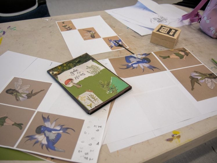 Image of A table displays printed illustrations of fairies, a sketch, a green book titled A Style for the Fairies, a pink bag, and a block stamp scattered on its surface.
