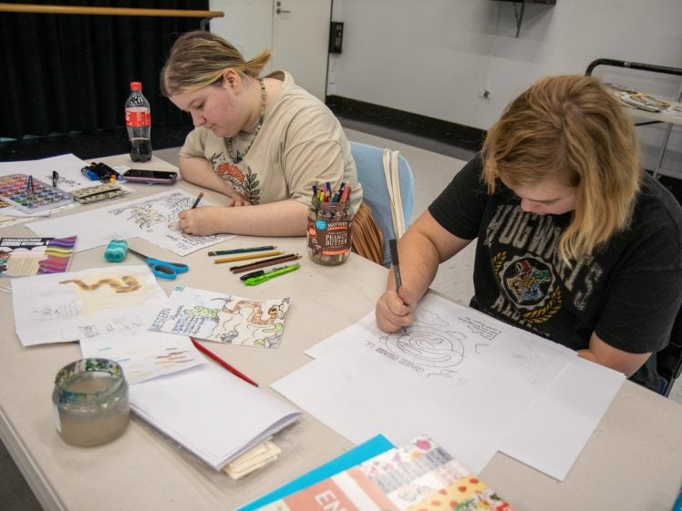 Image of Two people sit at a table drawing with pencils and coloured markers. Art supplies, paper, and coloured pencils are spread around them, and various sketches and art materials are visible on the table.