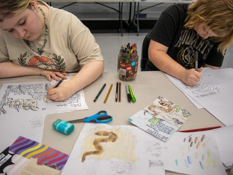 Image of Two kids sit at a table drawing and colouring on paper with felt-tip pens and coloured pencils. Art supplies, sketches, and a jar of coloured pencils are scattered on the table. Both kids are focused on their artwork.