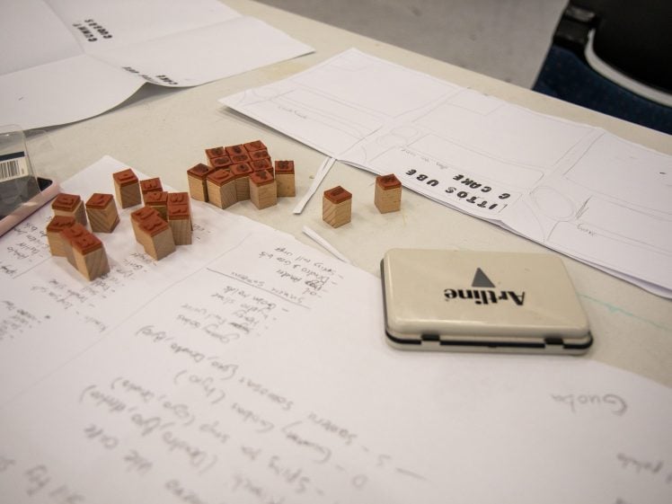 Image of A desk with scattered letter stamps, ink pad, handwritten notes, and paper templates labelled Little UBC Camp. Some stamps are grouped together; papers cover most of the surface.