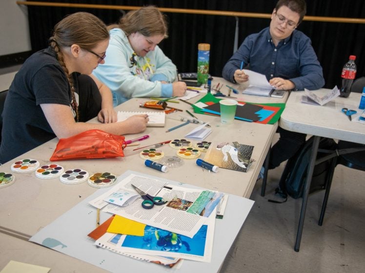Image of Three people sit at tables working on art projects with paper, scissors, watercolours, and felt-tip pens. Art supplies and magazine cut-outs are spread out across the tables in a creative, collaborative setting.