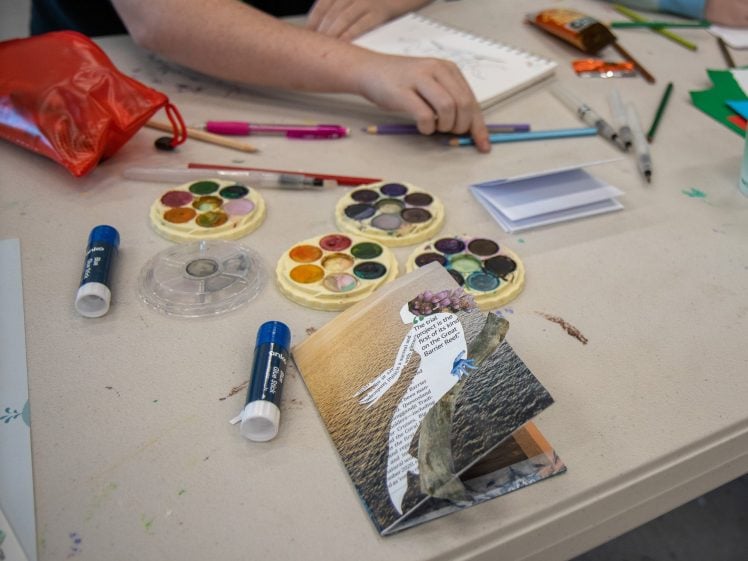 Image of A person sits at a table with watercolour paints, glue sticks, pens, a notebook, and a red pouch. In front of them is a handmade collage card featuring a cut-out figure and text.