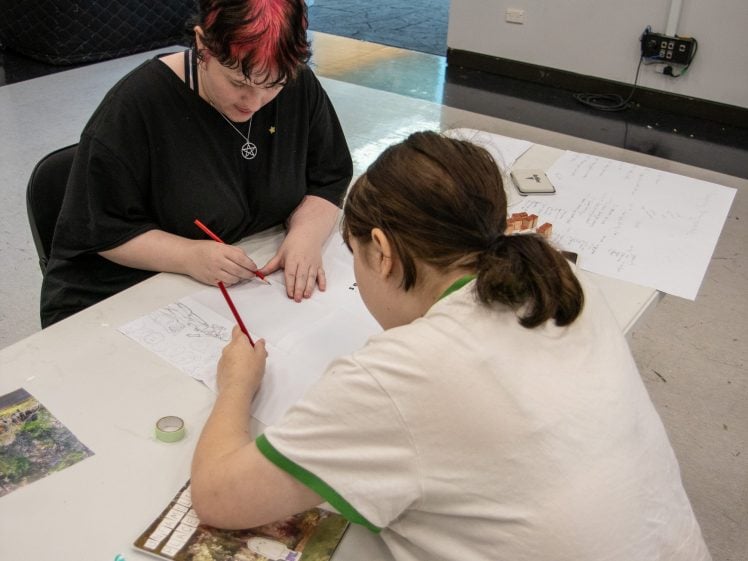 Image of Two people sit at a white table drawing or working on art projects with pencils and paper. One person has short black and red hair, and the other has brown hair tied back. Art supplies and papers are scattered on the table.