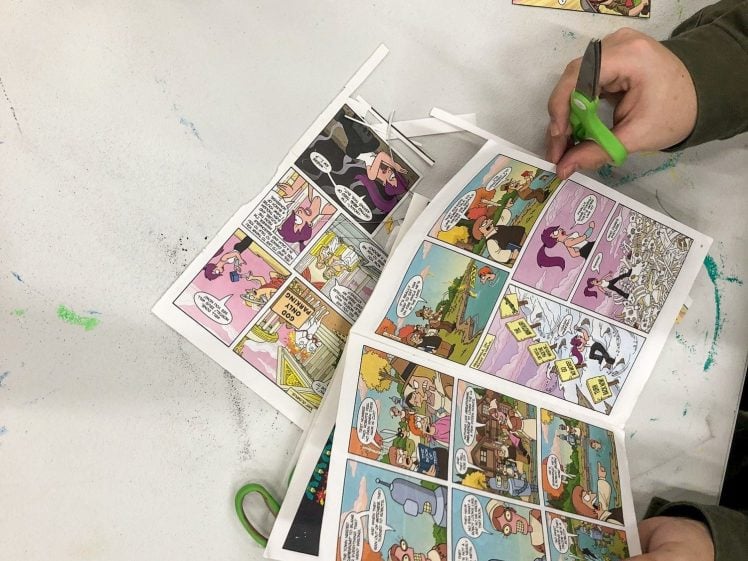 Image of A person uses green scissors to cut out colourful comic strips from printed sheets on a white table, surrounded by glue, cut-out comics, and crafting supplies.