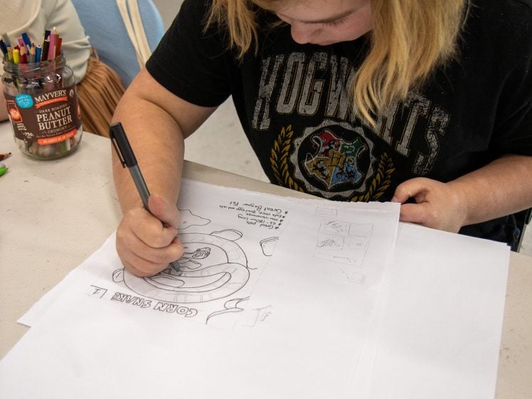 Image of A person with blonde hair wearing a Hogwarts T-shirt draws and writes on white paper at a table covered with art supplies, including coloured markers and a jar. Their focus is on a sketch in front of them.