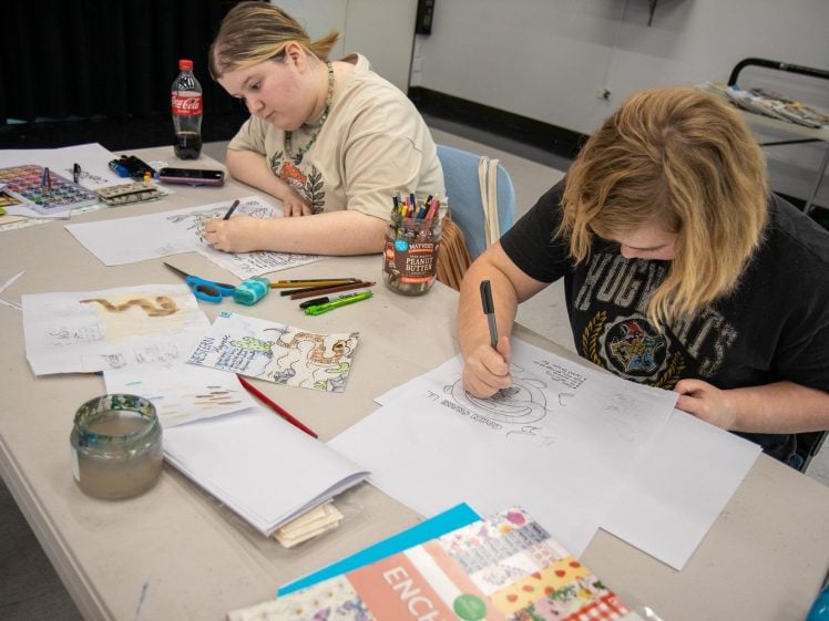Image of Two people sit at a table drawing on paper with various art supplies scattered around, including coloured markers, watercolour paints, sketchbooks, and a bottle of fizzy drink. Both appear focussed on their artwork.