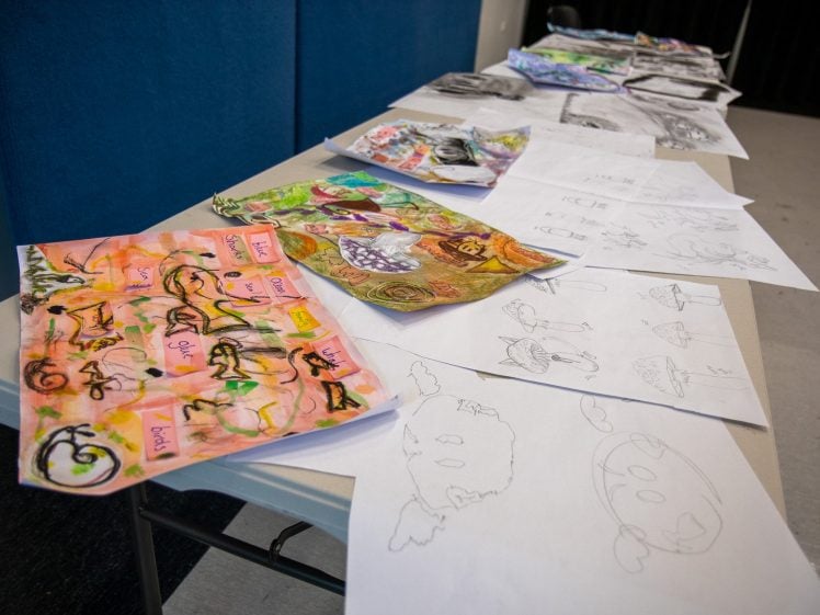 Image of A table covered with various colourful and black-and-white drawings and sketches, including abstract designs, cartoonish faces, and imaginative figures, arranged in a row in a classroom or art studio setting.