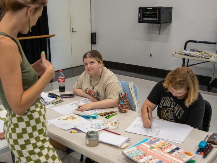 Image of Two people sit at a table drawing and colouring with art supplies, while a third person stands talking to them. Art books, coloured pencils, and a fizzy drink bottle are on the table in a classroom setting.