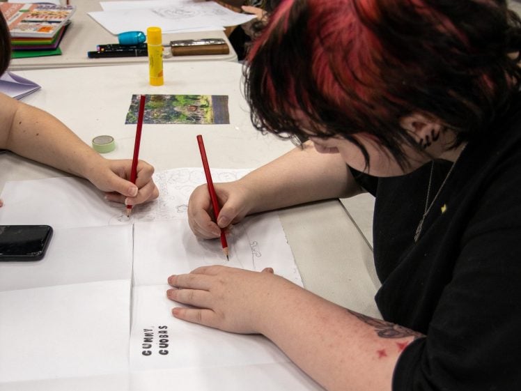 Image of A person with short black and red hair draws with a red pencil on a folded sheet of paper labelled CAKE COMICS and LET'S USE CAKE at a table with art supplies and other people working.