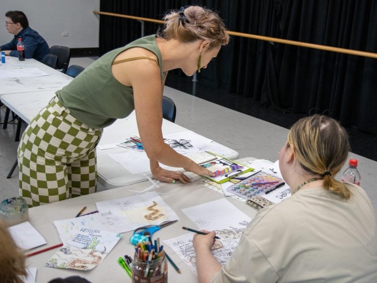 Image of A woman in a green top and chequered trousers leans over a table, assisting a seated person with colourful artwork. Other people are drawing at the table, which is covered with art supplies and sketches.