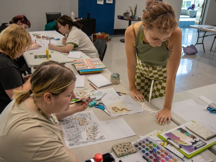 Image of Several people sit at tables drawing, painting, and colouring with art supplies, while a woman in green chequered trousers stands and looks at one person's artwork in a bright, creative studio space.