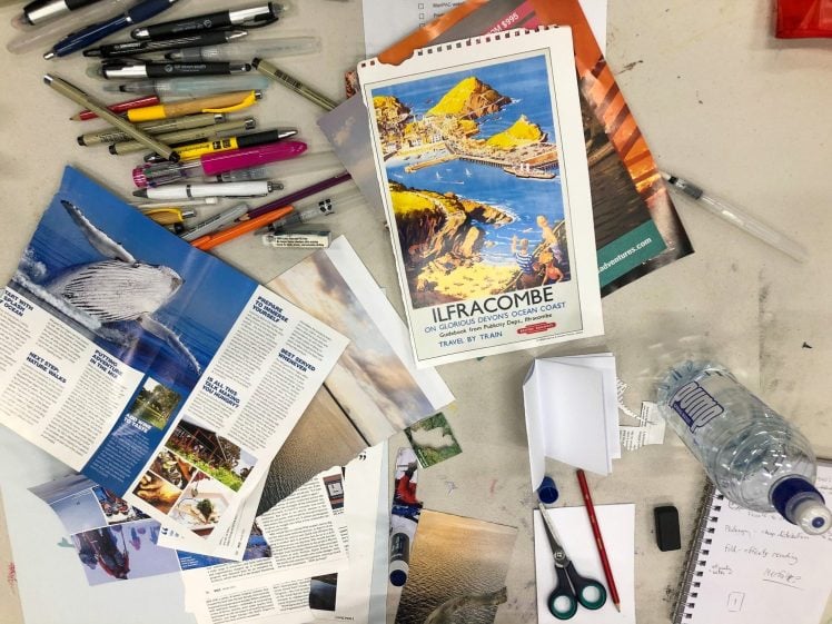Image of A cluttered desk with brochures, pens, coloured markers, scissors, a water bottle, and a vintage Ilfracombe travel poster, surrounded by scattered papers and a handwritten notepad.