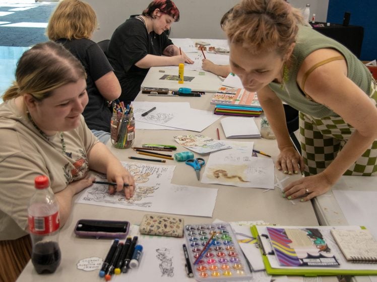 Image of Four people sit and work on drawings at a table covered with art supplies, paper, and drinks. One person leans over to engage with another, creating a collaborative and creative atmosphere.