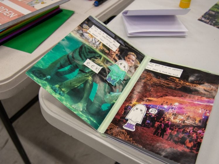 Image of An open scrapbook on a table shows colourful collages, including cut-out photos, a ghost illustration, and text like eek! and Ghost goes to a dear concert…. Other art supplies and papers are nearby.