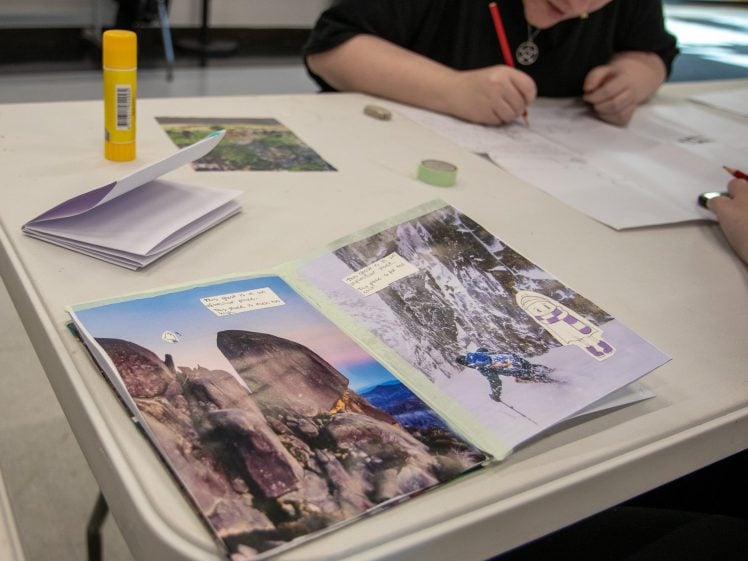 Image of A person sits at a table, drawing on paper. On the table are a glue stick, washi tape, a small notebook, and a collage book with mountain photos and cut-out characters.