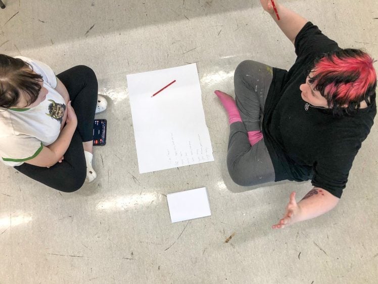 Image of Two people sit on the floor facing each other with a blank sheet of paper and a red pencil between them, appearing to discuss or brainstorm ideas. One gestures with their hand whilst the other listens.