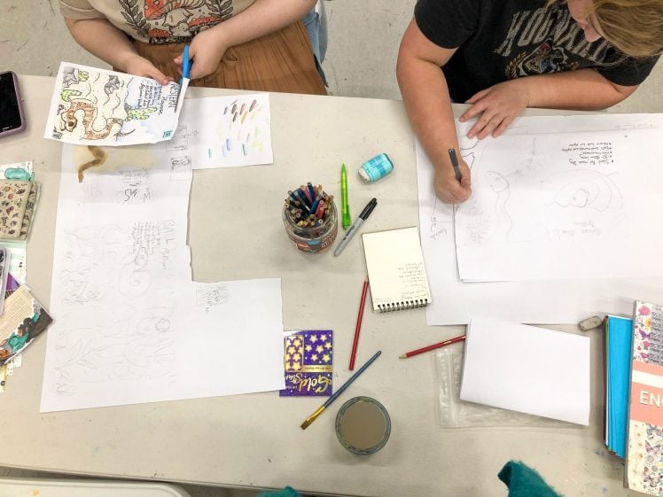 Image of Two people sit at a table drawing on white paper with pencils and coloured pens. Art supplies, coloured pencils, rubbers, a mug, and scattered papers are visible on the tabletop.