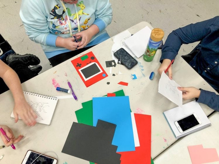 Image of Three people sit at a table covered with coloured paper, notebooks, pens, and mobile phones, working on arts and crafts projects. One person holds scissors, another sketches, and a third reaches for a pen.