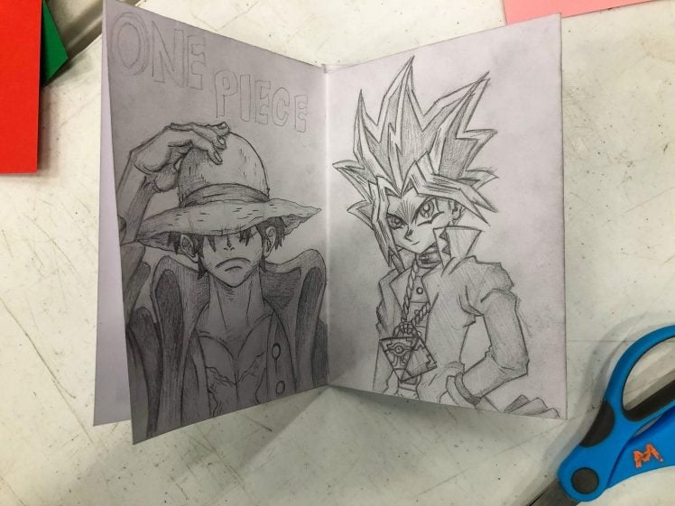 Image of An open sketchbook shows pencil drawings of Luffy from One Piece on the left and Yugi from Yu-Gi-Oh! on the right. The title ONE PIECE is written above Luffy. Blue scissors lie nearby on a white surface.