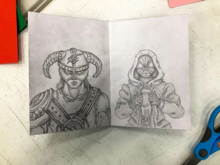 Image of A pencil drawing on folded paper shows two characters: one wearing a horned helmet and armour, and the other in a hooded jacket with a mask and glasses. The drawing is placed on a white table near scissors.