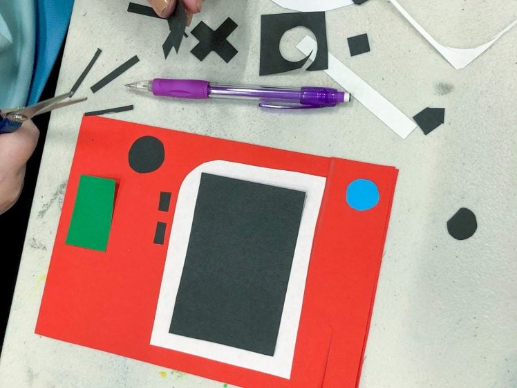 Image of A person uses scissors to cut black paper shapes for a red paper craft project. The table also has a purple pen, a mobile, and scattered paper scraps. The craft includes various coloured paper shapes.