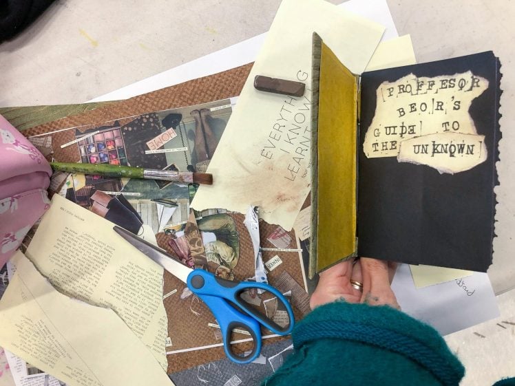 Image of A person holds an open handmade book titled Professor Beor's Guide to the Unknown above a table with scissors, pencils, paintbrush, torn book pages, and craft supplies scattered about.