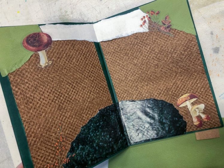 Image of An open handmade journal with textured brown and green pages, decorated with cut-out images of mushrooms, moss, and small plants along the top and bottom edges. The background surface is speckled and paint-stained.