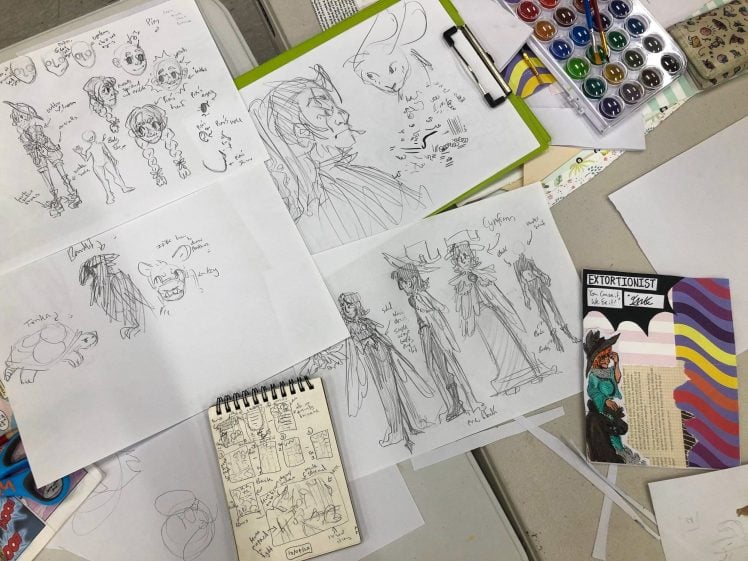 Image of A workspace with several character sketch sheets, a small comic notebook, a box of coloured pencils, watercolour paints, and a colourful zine titled EXTORTIONIST laid out on a table.