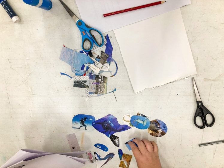 Image of A hand arranges cut-out images and shapes on a white table, surrounded by scissors, a red pencil, glue stick, and a blank sheet of paper. The cut-outs appear colourful, mostly in blue tones.
