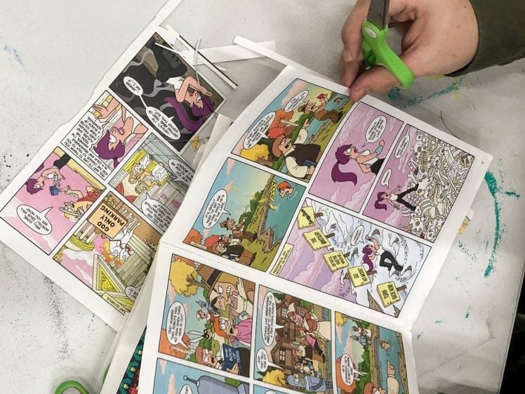 Image of A person uses green-handled scissors to cut out panels from a colourful comic book while seated at a white table. Another pair of scissors and comic pieces are visible on the table.