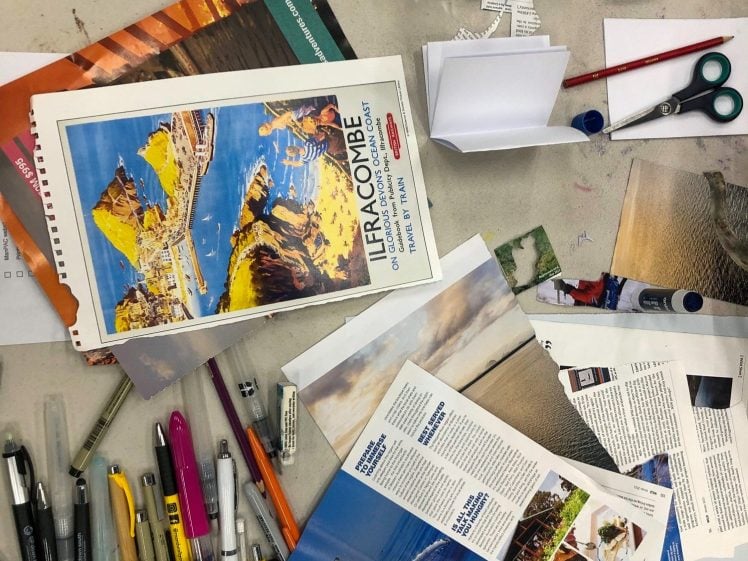 Image of A cluttered workspace with pens, markers, scissors, a water bottle, notebooks, and travel brochures featuring scenic images and a whale. Papers and leaflets are spread out across the table.