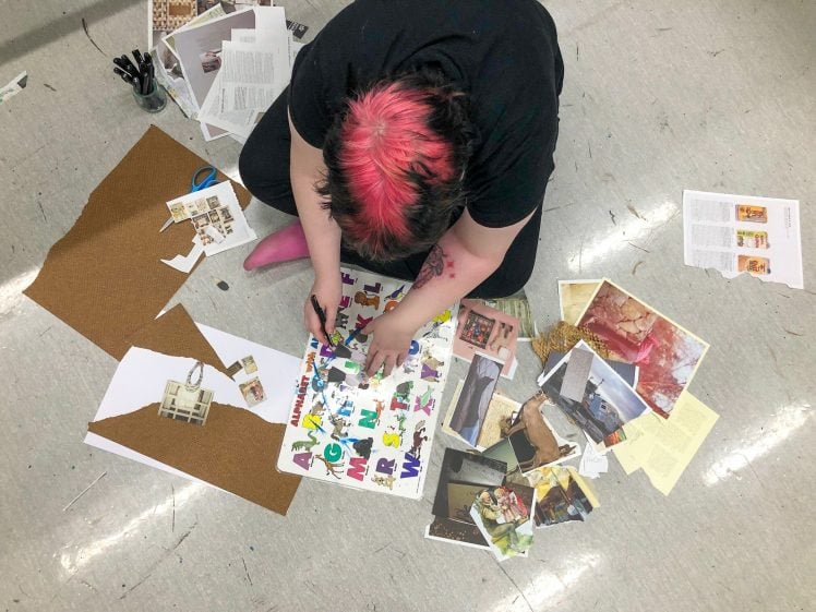Image of A person with short hair dyed black and pink sits on the floor, creating a collage with cut-out letters, magazine pages, and photos scattered around them on a grey tile floor.