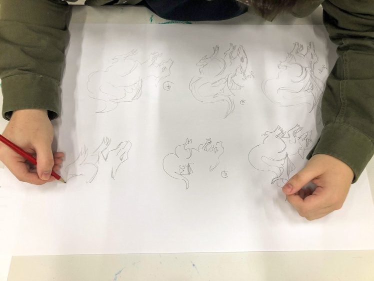 Image of A person wearing a green long-sleeved shirt is drawing sketches of animals, possibly dinosaurs, on a large white sheet of paper using a red pencil. Multiple unfinished sketches are visible.