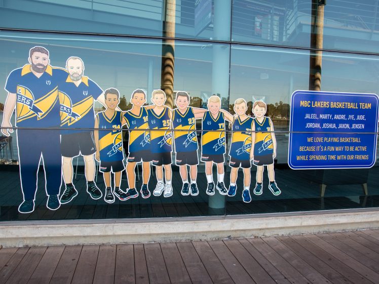 Image of Cartoon drawings of a basketball coach and seven children in blue and yellow uniforms are displayed on a glass wall. A sign lists their names and mentions they enjoy playing basketball and spending time with friends.