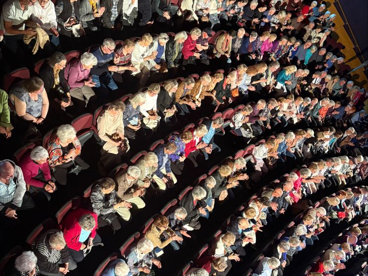 Image of A large audience sits in curved rows inside a theatre, viewed from above. Most seats are filled, and the people appear to be waiting for a performance or event to begin.