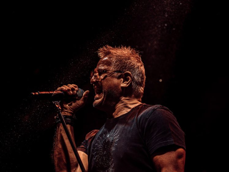 Image of A man wearing glasses passionately sings into a microphone on stage, illuminated by a dramatic spotlight against a dark background. Dust or mist particles are visible in the light.