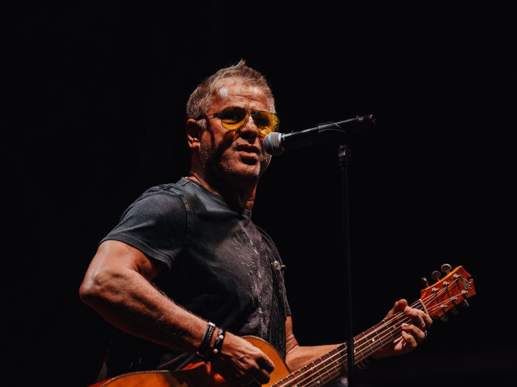 Image of A male musician wearing sunglasses plays an acoustic guitar and sings into a microphone on a dark stage, illuminated by spotlight.