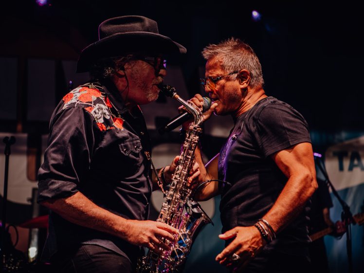 Image of Two male musicians perform passionately on stage; one plays the saxophone and wears a black hat and shirt with red patterns, whilst the other, wearing glasses and a black T-shirt, sings closely into the saxophone’s microphone.
