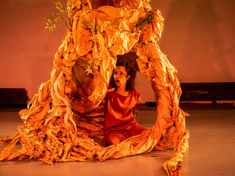 Image of A person in an orange outfit sits on the floor, partially surrounded by a large, crumpled, orange-brown paper structure resembling tree roots, with a surprised expression. The background is warmly lit.