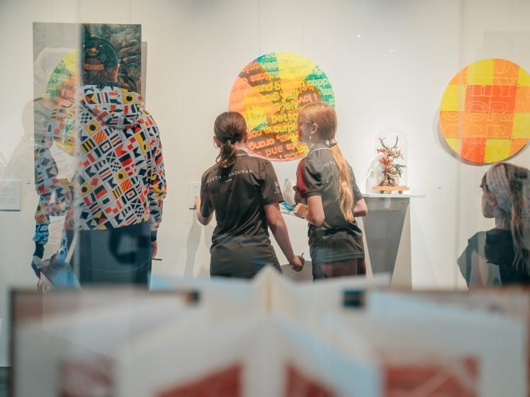 Image of Two girls stand hand in hand, viewing colourful circular artworks in a gallery. Other people are nearby, some partially visible. The scene has reflections on the glass, and open books are visible in the foreground.