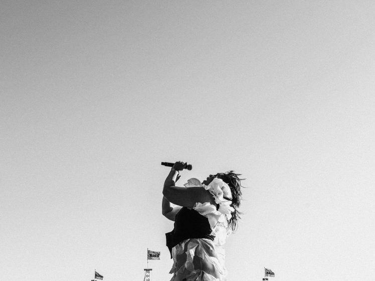 Image of A singer in a dramatic dress performs passionately on an outdoor stage in front of a large crowd under a clear sky, captured in black and white. Stage speakers and flags are visible in the background.
