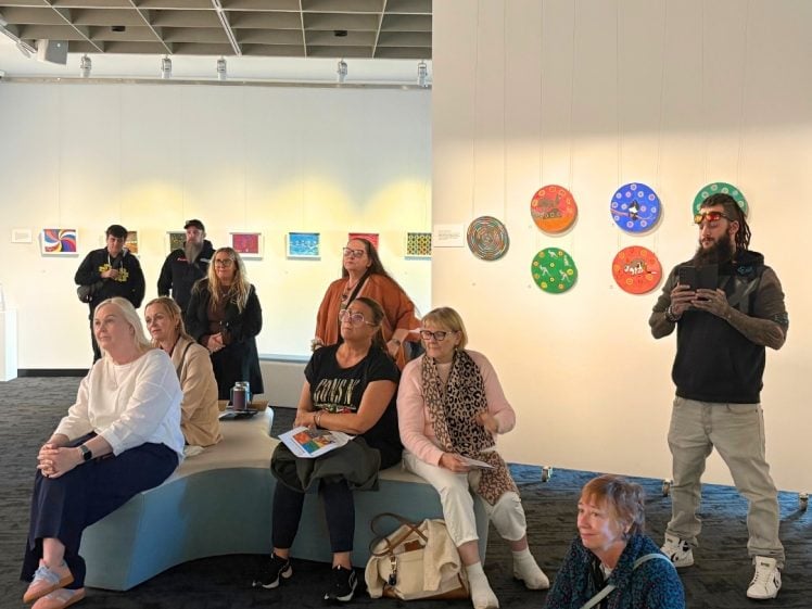 Image of A group of people sit and stand in an art gallery, attentively watching something off-camera. Colourful artwork is displayed on the white walls behind them under bright lighting.