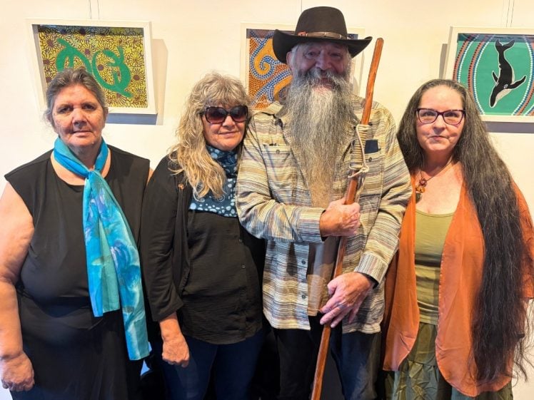 Image of Four adults stand together indoors, smiling for the camera. Three women and one man with a long white beard and hat, holding a wooden staff. Abstract art pieces are displayed on the wall behind them.